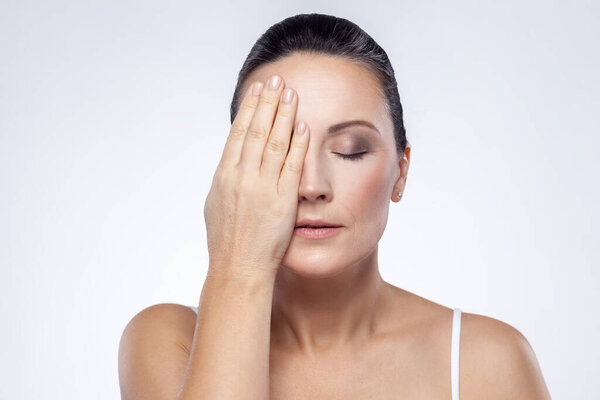 Closeup portrait of middle aged woman with perfect face skin hiding one eye with hand, keeps eyes closed, showing result of lifting procedures. Indoor studio shot isolated on white background.