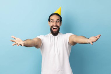 Beyaz tişört ve parti konisi giyen sakallı, şaşırmış bir adamın portresi, doğum günü partisini kutlayan, konukları davet eden, mutluluğu ifade eden. Mavi arka planda kapalı stüdyo çekimi.