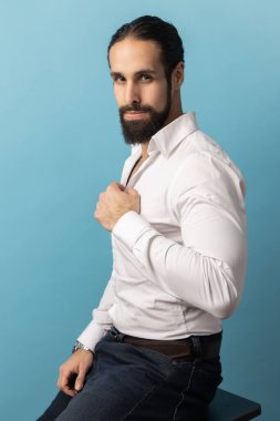 Sakallı, beyaz tişörtlü, siyah pantolonlu, gizemli bir şekilde kameraya bakan güçlü, kendine güvenen bir adamın portresi. Mavi arka planda kapalı stüdyo çekimi.