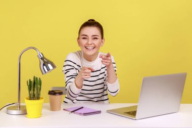Çizgili tişört giyen iyimser kadın ofis müdürü kamerayı gösteriyor ve samimi bir şekilde gülüyor, iş yerinde ev ofisinde oturuyor. Sarı arka planda kapalı stüdyo çekimleri.