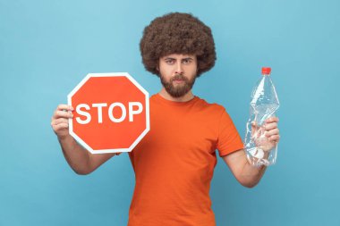 Ciddi sorumluluk sahibi, Afro saçlı, turuncu tişörtlü kırmızı dur işareti taşıyan ve kameraya bakan bir adamın portresi. Mavi arka planda kapalı stüdyo çekimi.