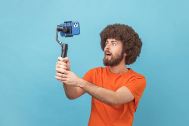 Afro saçlı, turuncu tişörtlü, büyük gözlü ve selfie çekmek için sabit duran şaşırmış bir adamın portresi. Mavi arka planda kapalı stüdyo çekimi.