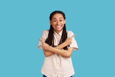 Son derece mutlu, siyah rastalı bir kadın kollarını çaprazlıyor ve farklı taraflarda beyaz gömlek giyen iki farklı nesne ya da varyant gösteriyor. Mavi arka planda kapalı stüdyo çekimi.