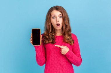 Şaşırmış bir kadın ağzı açık pembe kazak giymiş, elinde reklamcı için boş bir ekran olan akıllı bir telefonu tutuyor. Mavi arka planda kapalı stüdyo çekimi.