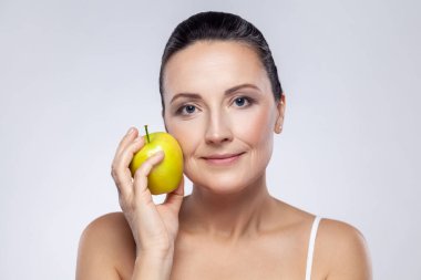 Yeşil elma tutan orta yaşlı güzel bir kadının yakın plan portresi. Meyve yemeyi seviyor. Sağlığını, cildini, kilo kontrolünü sağlıyor. Gri arka planda izole edilmiş kapalı stüdyo görüntüleri.