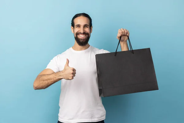 Portrait of man with beard wearing white T-shirt showing thumbs up holding shopping bag, pleased with good shopping and discounts, recommending. Indoor studio shot isolated on blue background.