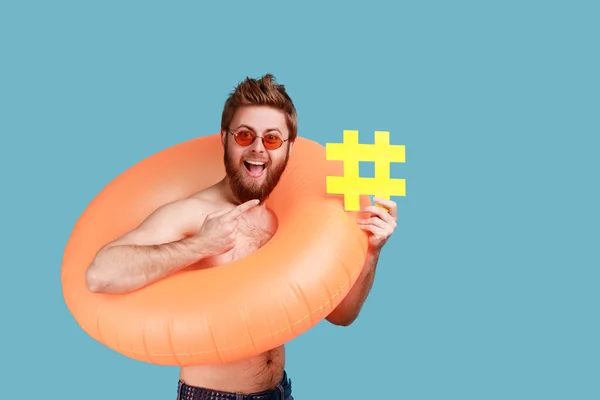 一个快乐的大胡子男人站在橙色的橡胶圈里 指着黄色的标签符号 是病毒网络思想的散列符号 室内拍摄被蓝色背景隔离 — 图库照片