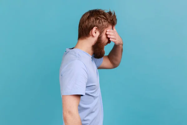 Don\'t want to look at this. Side view of bearded man covering eyes and turning with disgust from something shameful, scared afraid to see. Indoor studio shot isolated on blue background.
