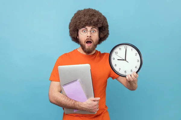 オレンジ色のTシャツを着たアフロの髪型をしたショックを受けた男の肖像画は 大きな壁の時計 締め切りを示す紙のノートパソコンを保持します 青い背景に隔離された屋内スタジオショット — ストック写真