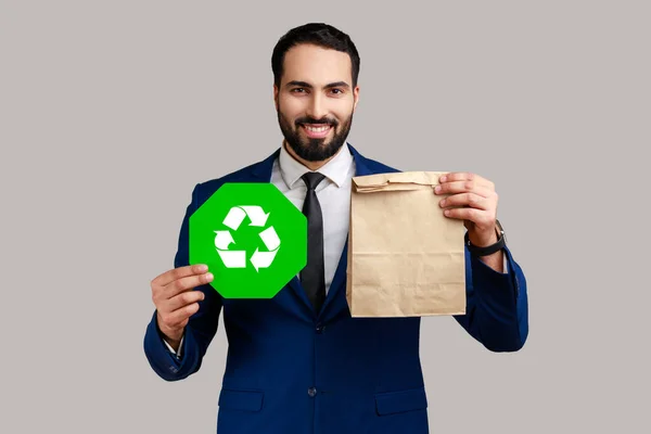 Portrait of smiling bearded man holding green recycling sign in hand and paper package, thinking green, wearing official style suit. Indoor studio shot isolated on gray background.