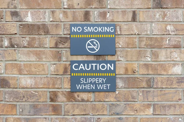 Warning signs No smoking and Slippery when wet on brick wall, warning labels on gray plates.