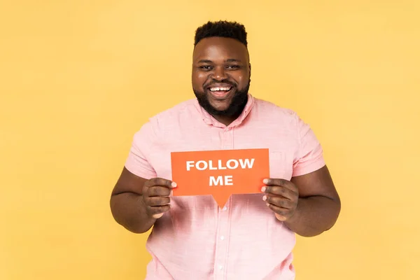 Portrait of happy optimistic man blogger wearing pink shirt standing holding card with follow me inscription, looking at camera with toothy smile. Indoor studio shot isolated on yellow background.