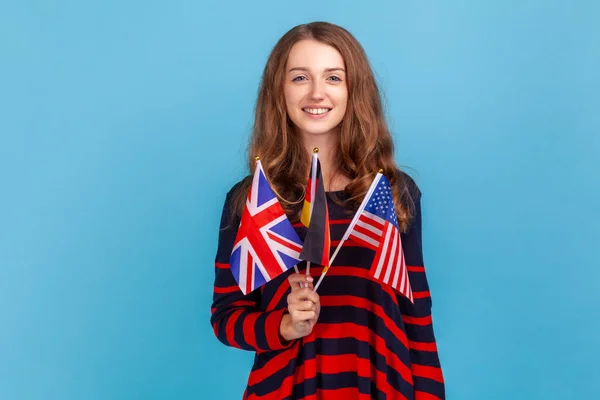 年轻成年女性穿着条纹休闲装的肖像 展示英国 美国和德国国旗 表达快乐 室内拍摄被蓝色背景隔离 — 图库照片