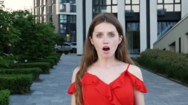 Şaşırmış, uzun saçlı, kırmızı şık elbiseli, şok olmuş yüzüyle kameraya bakan, büyük gözlü ve ağzı açık bir kadın. Arka planda modern bina ile açık hava çekimi.
