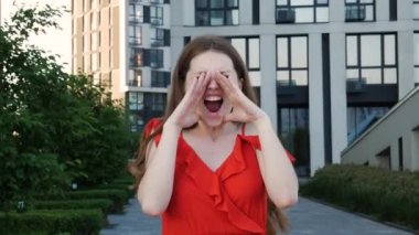 Uzun saçlı, kırmızı şık elbiseli kızgın bir kadın ayakta durup kötü haberler hakkında bağırıyor ya da ellerini ağzında tutmakta zorlanıyor. Arka planda modern bina ile açık hava çekimi.
