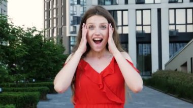 Kırmızı zarif elbiseler giyen, ağzı açık duran, hoş bir şaşkınlık içinde, şok edici bir ifade takınan, inanılmaz haberler duyan, ellerini kaldıran bir kadın. Arka planda modern bina ile açık hava çekimi.