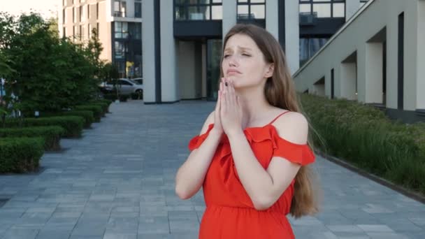 頼むお願いだ 赤いエレガントなドレスを身に着けている不幸な女性は 祈りの中で手を維持表情を暗示し 誠実な許可を求めて探します 現代的な建物を背景に屋外ショット — ストック動画