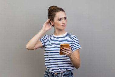 Çizgili tişört giyen düşünceli bir kadının portresi cep telefonuyla dikiliyor, kafaya dokunuyor, şüphe uyandırıyor. Gri arka planda izole edilmiş kapalı stüdyo görüntüleri.