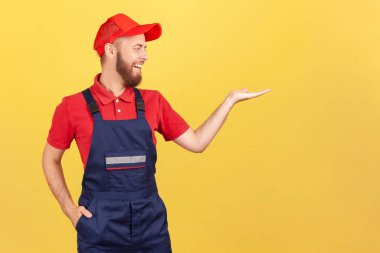 Gülümseyen bir işçinin portresi avucundaki reklam için fotokopi alanını sunuyor, başka yere bakıyor, tulum giyiyor ve sarı arka planda izole edilmiş kırmızı şapkalı stüdyo resmi var..