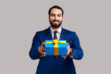 İyi görünümlü sakallı iş adamı mavi paket hediye kutusu veriyor, bayramlarla kutluyor, hediyesini tutuyor, resmi tarz takım elbise giyiyor. Gri arka planda izole edilmiş kapalı stüdyo görüntüleri.