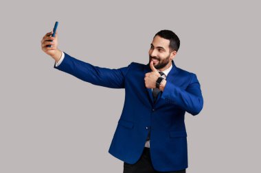 Akıllı telefon kamerasıyla selfie çeken olumlu bir adam, blogcu iletişimi, takipçiler için video kaydı, baş parmağını gösterme, resmi tarz takım elbise giyme. Gri arka planda izole edilmiş kapalı stüdyo görüntüleri.