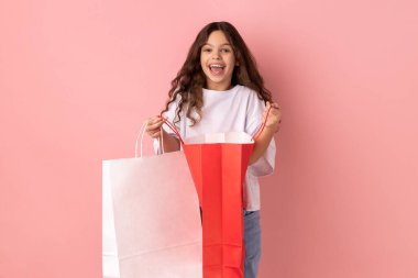 Beyaz tişörtlü, mutlu bir ifadeyle kameraya bakan ve elinde alışveriş torbaları tutan heyecanlı küçük kızın portresi. Pembe arka planda kapalı stüdyo çekimleri.