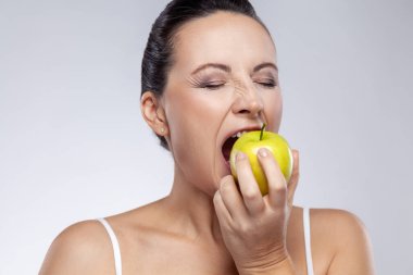 Taze yeşil elma ısıran orta yaşlı kadın portresi, doğal kozmetik, sağlıklı beslenme, sağlık ve cilt bakımı için vitaminler kullanıyor. Gri arka planda izole edilmiş kapalı stüdyo görüntüleri.
