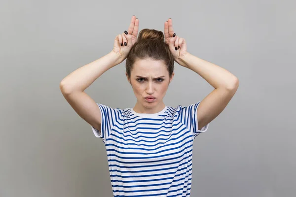 Portrait of angry bully woman wearing T-shirt showing bull horns with fingers on head, antler gesture, threatening with hostile view, ready to attack. Indoor studio shot isolated on gray background.