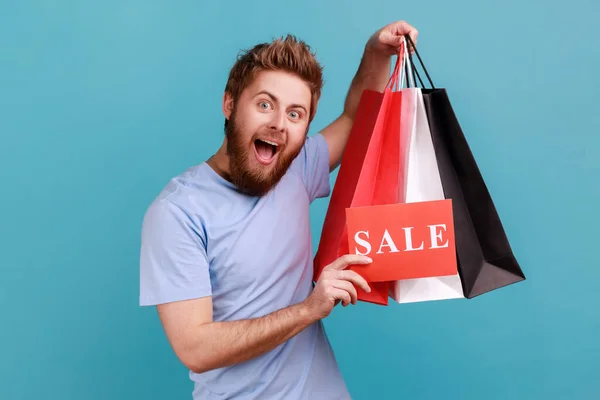 Portrait of handsome bearded man holding paper bags and sale sign, shopping and discounts, looking at camera with excited expression. Indoor studio shot isolated on blue background.