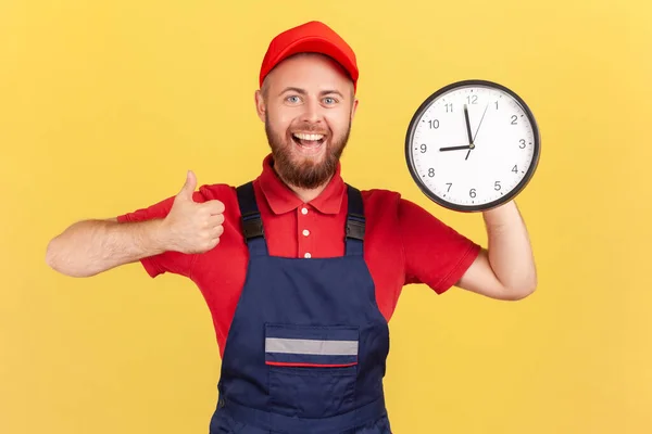 Portrait of happy smiling handyman standing with big wall clock in his hands, looking at camera and showing thumb up, wearing overalls and red cap. Indoor studio shot isolated on yellow background.