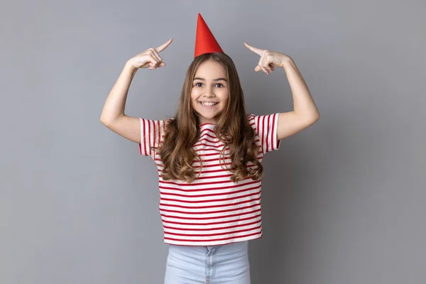 縞模様のTシャツを着た少女が頭の上にパーティーコーンを指してカメラを見て笑顔で誕生日を祝う姿が描かれています 灰色の背景に隔離された屋内スタジオショット — ストック写真