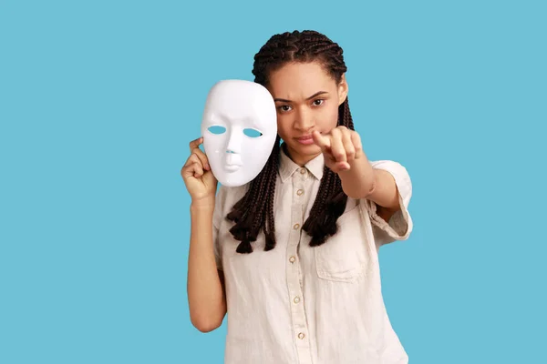 Strict bossy woman with black dreadlocks holding white mask in hand, pointing finger to camera with serious expression, wearing white shirt. Indoor studio shot isolated on blue background.