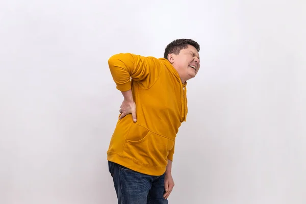 Spine problems. Middle aged man holding his back, having sudden lower back pain, pinched nerve or kidney inflammation, wearing urban style hoodie. Indoor studio shot isolated on white background.