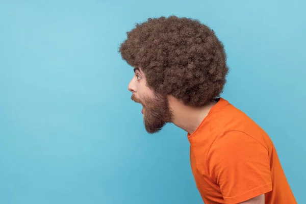 Side view of amazed man with Afro hairstyle wearing orange T-shirt standing with mouth open in surprise, has shocked expression, hears unbelievable news. Indoor studio shot isolated on blue background