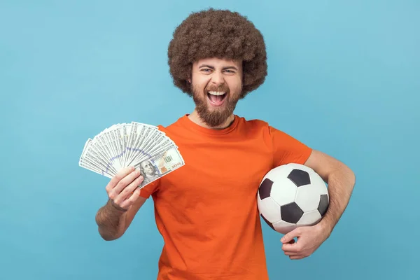 Portrait of happy man with Afro hairstyle wearing orange T-shirt holding soccer ball and hundred dollar bills, looking camera, betting and winning. Indoor studio shot isolated on blue background.