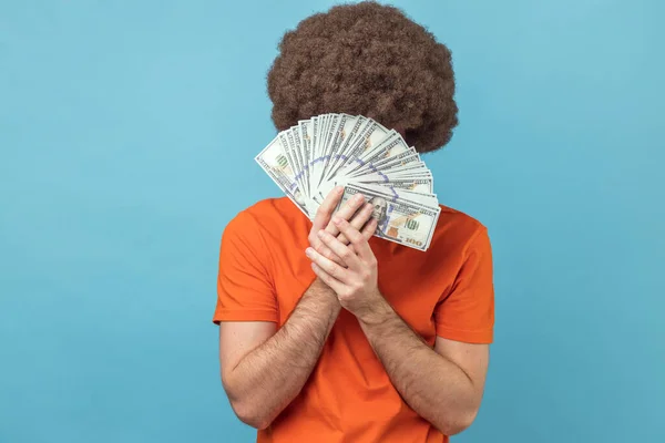Portrait of man with Afro hairstyle wearing orange T-shirt hiding face behind bunch of dollar banknotes, anonymous person holding money. Indoor studio shot isolated on blue background.