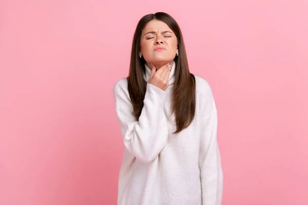 Portrait of sad ill woman touching her neck, suffering sore throat, viral infection or flu symptoms, wearing white casual style sweater. Indoor studio shot isolated on pink background.