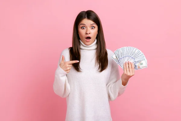 ドル紙幣のファンで衝撃的な笑みを浮かべて女性のポイントは 驚きの表情でカメラを見て 白いカジュアルなスタイルのセーターを着ている ピンクの背景に隔離された屋内スタジオショット — ストック写真