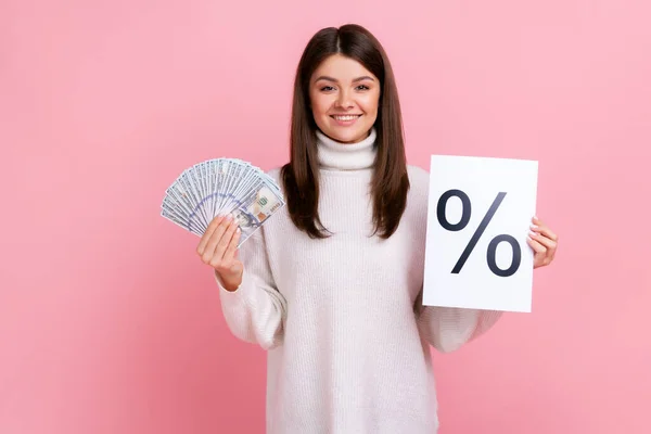 Smiling brunette female holding paper with percent mark and dollar banknotes, profitable bank offer, wearing white casual style sweater. Indoor studio shot isolated on pink background.