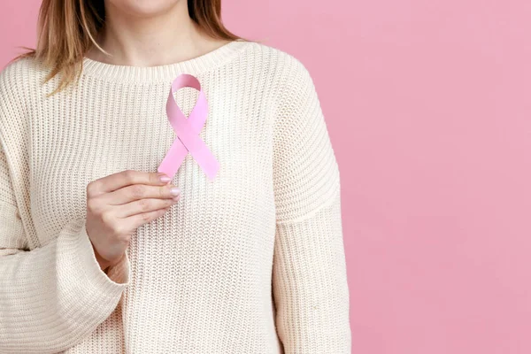 Female health check awareness. Closeup of woman hand in white pullover holding pink ribbon, symbol of breast cancer, timely diagnosis, support of oncology patients. indoor isolated on pink background