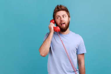 Tişörtlü, yakışıklı, sakallı bir adamın portresi. Retro telefondan kameraya konuşuyor, başka tarafa bakıyor, sohbet ediyor. Mavi arka planda kapalı stüdyo çekimi.