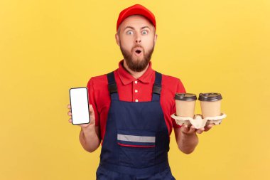 Mavi tulumlu, kırmızı şapkalı, elinde kahve tutan ve reklam için boş ekranlı cep telefonunu gösteren şok olmuş kurye adam. Sarı arka planda kapalı stüdyo çekimleri.