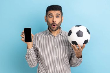 Futbol topu tutan sakallı adam ve akıllı telefon boş siyah ekran, basit bir şampiyonluk biletiyle şok olmuş, çizgili gömlek giymiş. Mavi arka planda kapalı stüdyo çekimi.