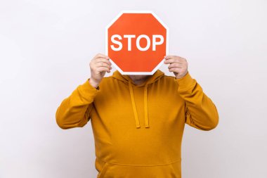 Dikkat, yasak yol. Bilinmeyen kişi yüzünü Dur sembolünün arkasına gizliyor, kırmızı trafik levhası uyarılmış, şehir tarzı kapüşonlu giyiyor. Beyaz arka planda izole edilmiş kapalı stüdyo görüntüleri.