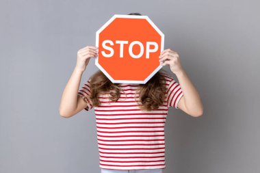 Çizgili tişörtlü, yüzünde dur işareti olan küçük bir kız kırmızı trafik levhası tutan, tehlike, kısıtlama ve sınırlama uyarısı yapan isimsiz bir kadın. Gri arka planda izole edilmiş kapalı stüdyo görüntüleri.