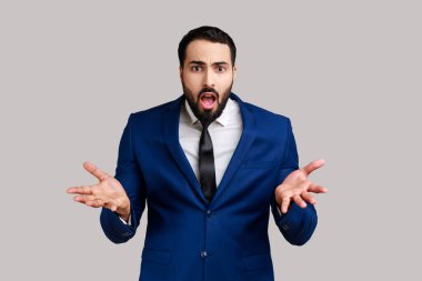 Elleri havada ve kızgın bir ifadeyle duran sakallı bir adamın portresi. Çatışma nedenini soruyor, resmi stil kıyafeti giyiyor. Gri arka planda izole edilmiş kapalı stüdyo görüntüleri.