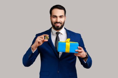 Gülümseyen pozitif sakallı iş adamı elinde mevcut kutu ve altın bitcoin ile duruyor. Şifreli para, resmi tarz takım elbise giyiyor. Gri arka planda izole edilmiş kapalı stüdyo görüntüleri.