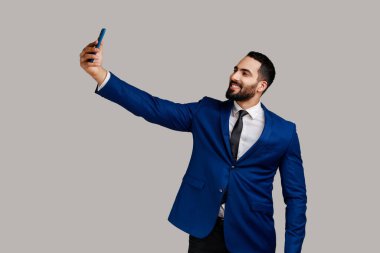 Sakallı adam akıllı telefon kamerasıyla selfie çekiyor, blogcu iletişimi kuruyor sosyal ağlardaki takipçiler için video kaydediyor, resmi tarz takım elbise giyiyor. Gri arka planda izole edilmiş kapalı stüdyo görüntüleri.