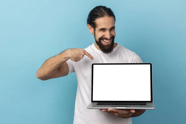 白いTシャツを着て髭を生やした男の肖像画は 空白の画面とカメラ インターネット広告で笑みを浮かべてノートパソコンを保持立っている 青い背景に隔離された屋内スタジオショット — ストック写真