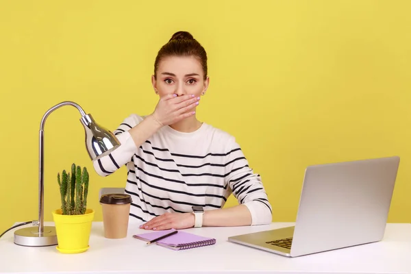 Portrait of shocked young woman office worker covering mouth, looking at camera with terrified expression, found scary secret. Indoor studio studio shot isolated on yellow background.